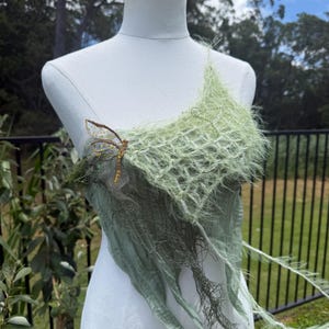 Può includere: Top verde chiaro fatto a mano con un ornamento a forma di libellula. Il top presenta un design a strati con uno scollo strutturato e sfrangiato e tessuto fluente. La libellula è dorata e ha un design con perline. Il top è esposto su un manichino.