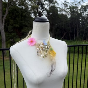 Pode incluir: Um colar decorativo com uma flor rosa, lantejoulas e penas em tons de amarelo, azul e cinza. O colar é exibido em um manequim branco. O fundo inclui uma cerca preta e folhagem verde.