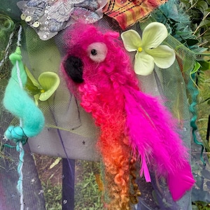 Pode incluir: Escultura de papagaio artesanal com lã rosa, laranja e amarela vibrante. O papagaio apresenta um bico preto, um olho branco e uma pena rosa brilhante. A escultura é adornada com flores de tecido e rede verde.