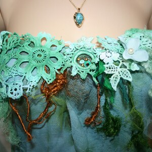 May include: A strapless top made of layers of green, blue and white lace, tulle and fabric. The top is decorated with a small, white flower with a pearl centre and a gold chain with a turquoise pendant.