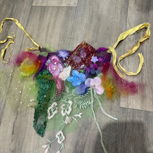 Pode incluir: Uma colorida coroa de fadas ou adorno de cabeça feito à mão. Apresenta uma mistura de tecidos, incluindo renda, tule e elementos bordados, adornado com flores artificiais, penas e fitas. A paleta de cores inclui tons de roxo, verde, azul e amarelo.
