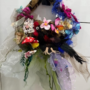 Op de afbeelding: Een handgemaakte decoratieve krans met een centrale vogel figuur met bruine en zwarte veren, versierd met kleurrijke vlinders, bloemen en tule. De basis van de krans is imitatiebont met een tijgerstreep patroon.