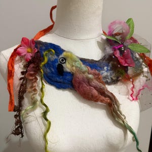 May include: A felted parrot-themed necklace with a blue base, adorned with colorful wool, pink flowers, and green leaves. The necklace features a black and yellow eye, and is embellished with orange ribbons and a sheer, pink-trimmed fabric.