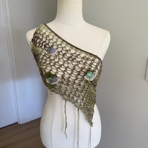 May include: An olive green, one-shoulder crochet top featuring an open, net-like stitch pattern. The garment is embellished with small, circular, sequined floral-like appliqués and a single central cowrie shell, with loose yarn ends hanging down.