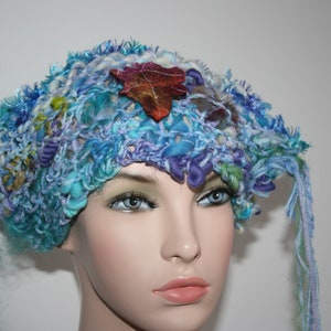 May include: A blue, purple, and white crocheted hat with a brown leaf accent. The hat has long, flowing strands of yarn.