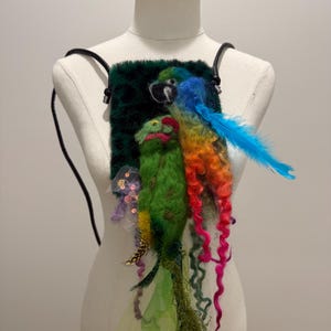 Pode incluir: Uma mochila artesanal e caprichosa com um design de papagaio vibrante. A bolsa tem um fundo verde escuro e texturizado com um papagaio feltrado em tons de azul, verde, laranja e vermelho, com uma longa pena azul. A bolsa tem alças pretas.