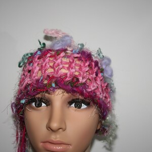 Może przedstawiać: Ręcznie robiona czapka w odcieniach różu, fioletu i zieleni. Czapka ma ażurowy wzór z błyszczącymi srebrnymi nićmi i ozdobnymi elementami. To wyjątkowe nakrycie głowy to stylowy dodatek.