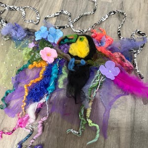 Pode incluir: Um acessório colorido e artesanal com tema de tucano, com uma gama vibrante de cores. O design incorpora um tucano amarelo e preto, rodeado por detalhes roxos, azuis, rosa e verdes, incluindo flores, penas e fios texturizados. O acessório tem tiras com padrões em preto e branco.
