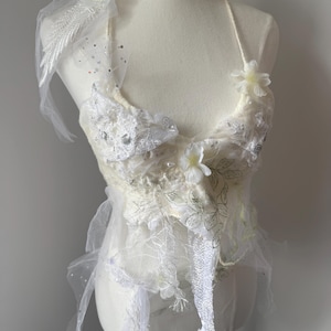 Puede incluir: Top hecho a mano de color marfil con detalles florales y de hojas. El top presenta delicados encajes, tul y adornos de lentejuelas. Un cuello halter está adornado con pequeñas flores blancas. El diseño incluye texturas en capas.