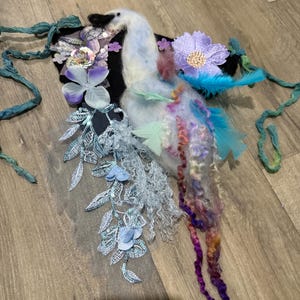 Pode incluir: Um cisne têxtil artesanal com bico e cabeça pretos, corpo branco e penas da cauda coloridas. O cisne é adornado com enfeites florais roxos e azuis, folhas verdes e fitas azuis. A obra de arte é colocada sobre um fundo preto.