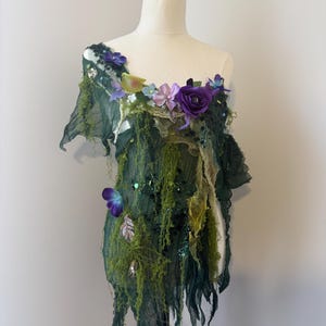 Puede incluir: Una parte superior de disfraz verde y cubierta de musgo, adornada con flores moradas y azules y una mariposa. La tela está drapeada y en capas, creando una apariencia caprichosa de hada. La parte superior se muestra en un maniquí.