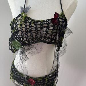 May include: Black crochet halter top and matching bottom set, adorned with faux greenery, butterflies, and flowers. The set is displayed on a mannequin. The top features a key charm and the bottom has a floral accent. The set is made of black crochet and lace.