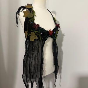 Può includere: Un top nero all'americana decorato con foglie, muschio e fiori rossi. Il top è adornato da un tessuto nero fluente. L'indumento è esposto su un manichino, mettendo in risalto il suo design e la sua consistenza unici.