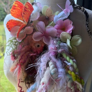 Pode incluir: Um colar artesanal com uma vibrante gama de cores e texturas. O design incorpora asas de borboleta laranja, flores rosa e brancas e uma mistura de fios e tecido. O colar é exibido em um manequim.