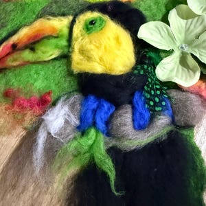 Pode incluir: Um tucano de feltro com cabeça amarela, corpo preto e um bico grande e colorido. A ave é cercada por feltro verde, azul e cinza, com alguns detalhes brancos e rosa. Uma flor verde clara no fundo.