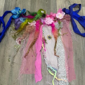 Pode incluir: Um traje de fada artesanal e caprichoso com uma criatura verde semelhante a um pássaro no centro. O traje apresenta tecidos rosa, azul e branco, adornados com flores, penas e fitas. O design inclui fitas longas e esvoaçantes e uma variedade de texturas.