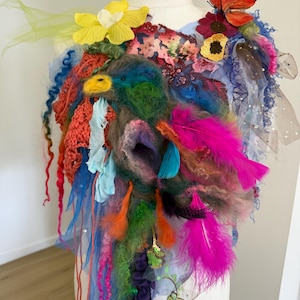 Pode incluir: Uma obra de arte têxtil colorida com uma variedade de texturas e embelezamentos. O design incorpora penas rosa brilhantes, flores amarelas e laranja e uma borboleta. Fios de tecido azul, laranja e roxo pendem da peça, criando um efeito em camadas.