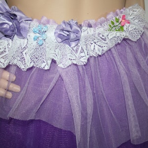 Könnte beinhalten: Ein lila Tüllrock mit weißer Spitzenborte und lila Satinblumen. Der Rock hat ein mehrschichtiges Design mit einer dunkleren lila Schicht darunter.