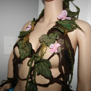 Può includere: Un capo fatto a mano, ispirato alla natura, con un top a forma di reggiseno e un pezzo per le spalle, adornato con foglie di feltro, viti e accenti floreali rosa. La tavolozza dei colori comprende sfumature di verde, marrone e rosa, creando un'estetica stravagante e organica.