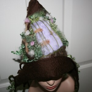 Puede incluir: Un sombrero de fieltro marrón con un diseño caprichoso con hilo y tela verde, morado y blanco. El sombrero tiene una ala ancha y una corona puntiaguda.
