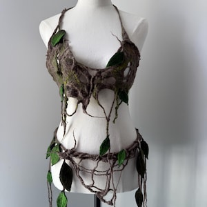 Può includere: Un completo di abbigliamento fatto a mano, ispirato alla natura, esposto su un manichino. Il set comprende un top all'americana e un pezzo in vita, entrambi realizzati in feltro marrone con accenti di foglie verdi. Il design evoca un tema forestale.