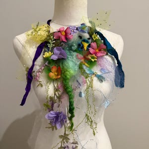 Op de afbeelding: Een bloemenhalsstuk met roze, paarse en gele bloemen, gepresenteerd op een mannequin. Het ontwerp bevat groene wol, transparante stof en blauwe en paarse linten. Het stuk heeft een grillige, artistieke uitstraling.
