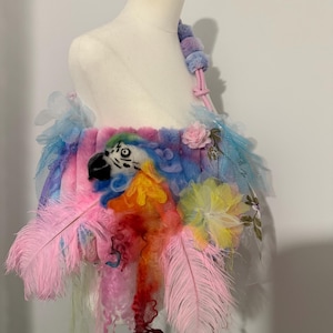 Op de afbeelding: Een kleurrijke, pluche, regenboogkleurige tas met een papegaaienkop en roze veren. De tas heeft een roze band en is versierd met bloemen en andere versieringen.