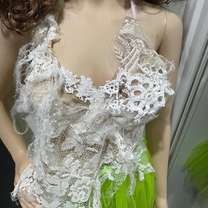May include: White lace halter top with a pink ribbon tie. The lace is frayed and distressed, giving it a vintage look. The top is layered over a bright green tulle skirt.