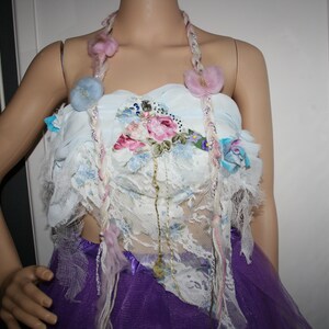 May include: A white lace top with floral embroidery and a purple skirt. The top has a pink and blue braided cord with flower accents.
