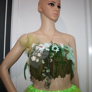 Puede incluir: Un top de encaje verde y blanco con adornos florales y un diseño de vid de cuentas. El top está puesto en un maniquí con un collar de oro.