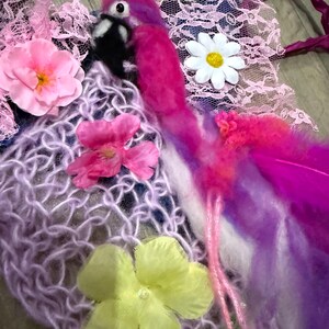 Pode incluir: Uma peça de arte têxtil artesanal com um flamingo rosa vibrante com bico e olho pretos. A obra de arte incorpora penas rosa e roxas, renda e flores de tecido em tons de rosa, branco e amarelo. O fundo inclui uma rede de malha.