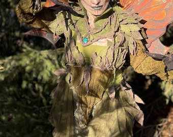 May include: A detailed costume of a forest creature with green and brown tones. The costume features a detailed face with horns, teal hair, and a green and gold outfit with leaf-like accents and large wings.