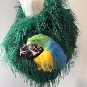 Op de afbeelding: Een groene schoudertas van veren met een vilten papegaaienkop. De papegaaienkop heeft blauwe, gele en groene veren, met een zwarte snavel en witte ogen. De tas wordt op een mannequin getoond.