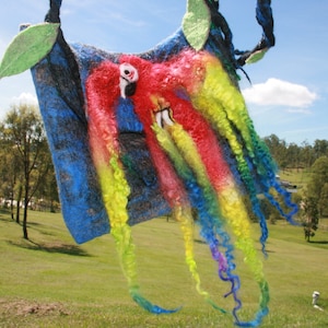 Op de afbeelding: Een kleurrijk wandkleed van vilt met een papegaai-ontwerp. De papegaai is rood, geel en blauw met lange, stromende garenstrengen in regenboogkleuren.