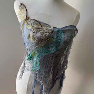 Puede incluir: Una prenda drapeada azul claro con una decoración de mariposa. La mariposa tiene detalles dorados y amarillos. La prenda está adornada con lentejuelas verdes y plateadas y una superposición en forma de red. La tela tiene una apariencia desgastada y etérea.