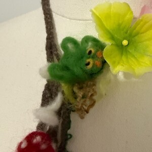 May include: A brown yarn necklace with a green felt creature, a red felt mushroom with white dots, and a yellow felt flower.