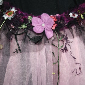 Könnte beinhalten: Ein rosa Tüllrock mit einem dekorativen Blumenband. Das Band hat eine lila Filzblume, gestickte Blumen und schwarze Tüllakzente. Der Rock ist mehrlagig und hat einen schimmernden Effekt, der einen skurrilen Look erzeugt.