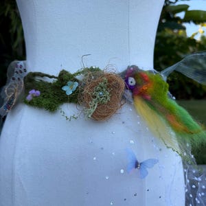 May include: A whimsical fairy belt, featuring a central design of moss, twigs, and small flowers. The belt has a colorful, feathered accent and a sheer, dotted skirt with a blue butterfly. The belt is displayed on a white mannequin.