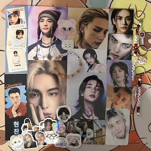 Peut inclure: Collection de produits dérivés Stray Kids, comprenant des cartes photo, des autocollants, un bracelet de perles avec le nom "HYUNJIN" et des porte-clés. Les articles présentent des images de l'artiste. La palette de couleurs comprend des bleus, des roses et des blancs.