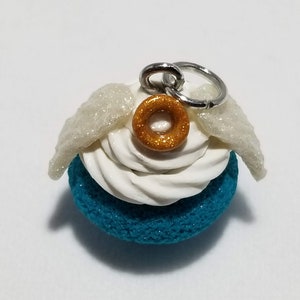 Peut inclure: Un cupcake bleu turquoise avec un gla&ccedil;age blanc et un beignet dor&eacute; paillet&eacute; sur le dessus. Le cupcake a des ailes blanches paillet&eacute;es et un charme argent&eacute; attach&eacute;.