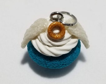May include: A teal blue cupcake with white frosting and a gold glitter donut on top. The cupcake has white glitter wings and a silver charm attached.