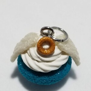 Peut inclure: Un macaron bleu turquoise avec un gla&ccedil;age blanc et un beignet dor&eacute; paillet&eacute; sur le dessus. Le macaron a des ailes blanches paillet&eacute;es attach&eacute;es sur le c&ocirc;t&eacute; et un anneau de saut argent&eacute; pour l'accrocher.