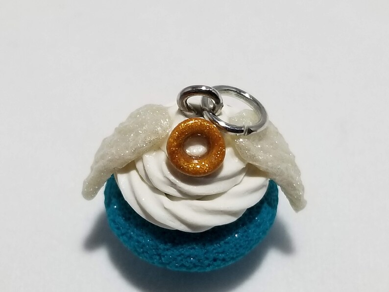 Peut inclure: Un macaron bleu turquoise avec un gla&ccedil;age blanc et un beignet dor&eacute; paillet&eacute; sur le dessus. Le macaron a des ailes blanches paillet&eacute;es attach&eacute;es sur le c&ocirc;t&eacute; et un anneau de saut argent&eacute; pour l'accrocher.