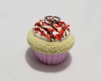 May include: A small, pink cupcake with white frosting, chocolate sauce, and red strawberry pieces. The cupcake has a silver charm attached to the top.