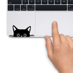 Op de afbeelding: Een zwarte kat silhouet sticker die over de rand van een laptop toetsenbord kijkt. De kat heeft grote ogen en een kleine neus.