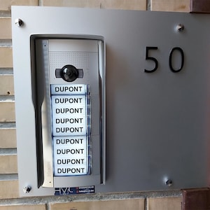 Può includere: Un pannello di interfono argentato con il numero 50 nero e un sistema di interfono blucino con il nome DUPONT ripetuto su otto pulsanti. Il sistema di interfono ha un piccolo obiettivo della telecamera e un'icona del telefono.