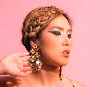 May include: A woman with a braided hairstyle and silver eyeshadow wears a sparkly, light purple one-shoulder top and large gold and black statement earrings.