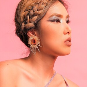 May include: A woman with a braided hairstyle and silver eyeshadow wears a silver sparkly top and large gold and orange statement earrings.