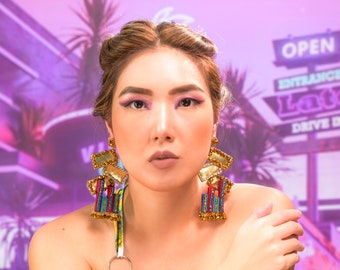 May include: A woman with brown hair and makeup is wearing large, colorful, gold-toned earrings. The earrings have a geometric design with red, blue, and green beads. The background is a blurred image of a pink and purple building with a neon sign that says "Late Drive In".