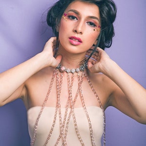 May include: A woman with dark hair and makeup featuring pink and gold glitter is wearing a delicate, multi-layered beaded necklace. The necklace is made of small, clear beads and has a silver clasp. The woman is wearing a strapless top and has her hands on her neck.